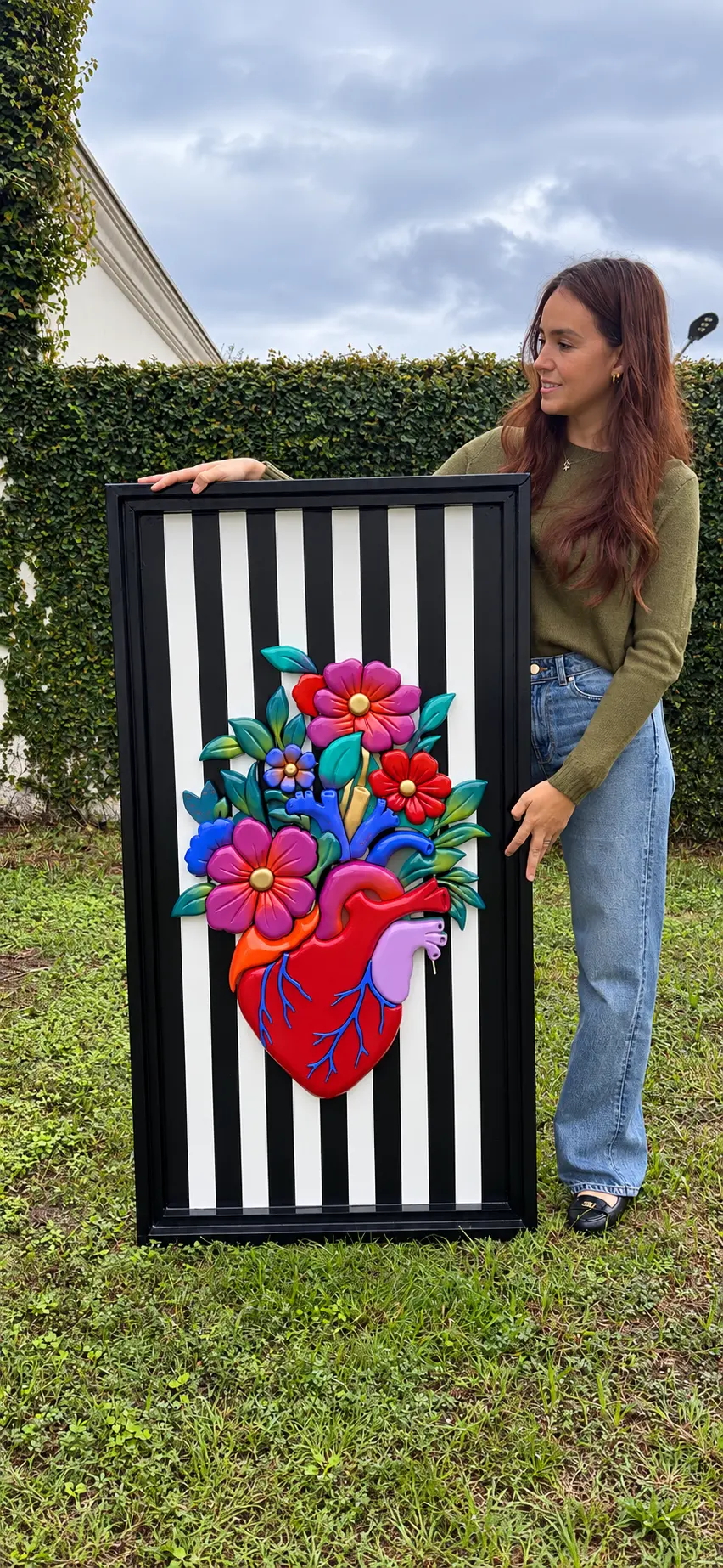 Renacer desde el corazón, obra de gran formato: corazón anatómico del que florece un jardín de flores sobre fondo de rayas verticales en blanco y negro, junto a una persona que muestra su escala